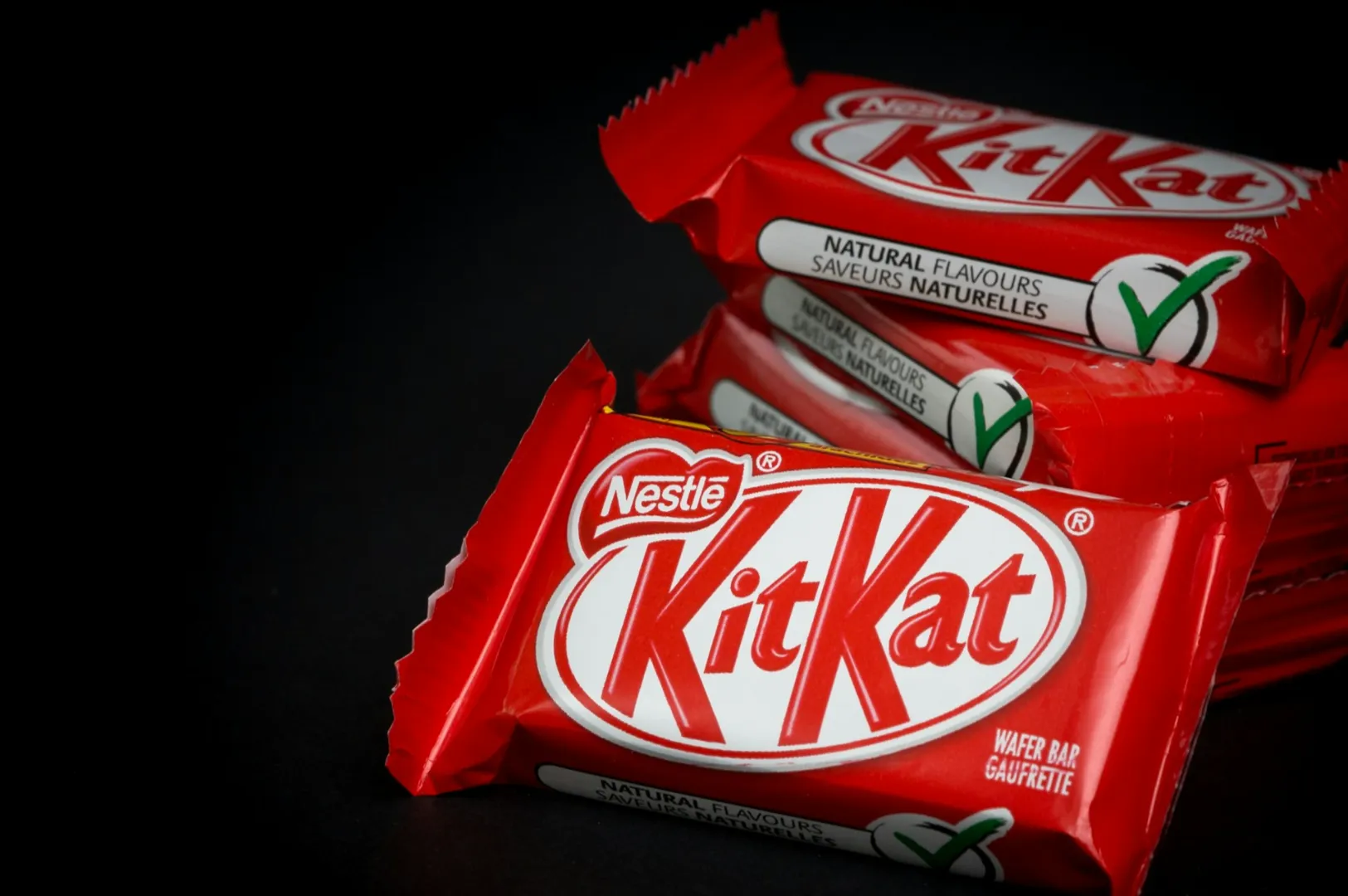 Red Kit Kat wrappers stacked against a black background, highlighting the white logo. Labels indicate natural flavors with a green checkmark.