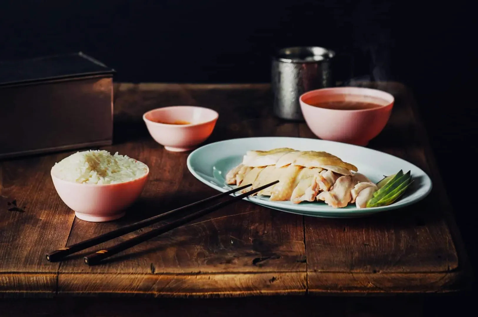 “A rustic wooden table set with a plate of sliced chicken and garnished greens, a bowl of white rice, a dipping sauce, and steaming soup, conveying a cozy, warm meal.”
