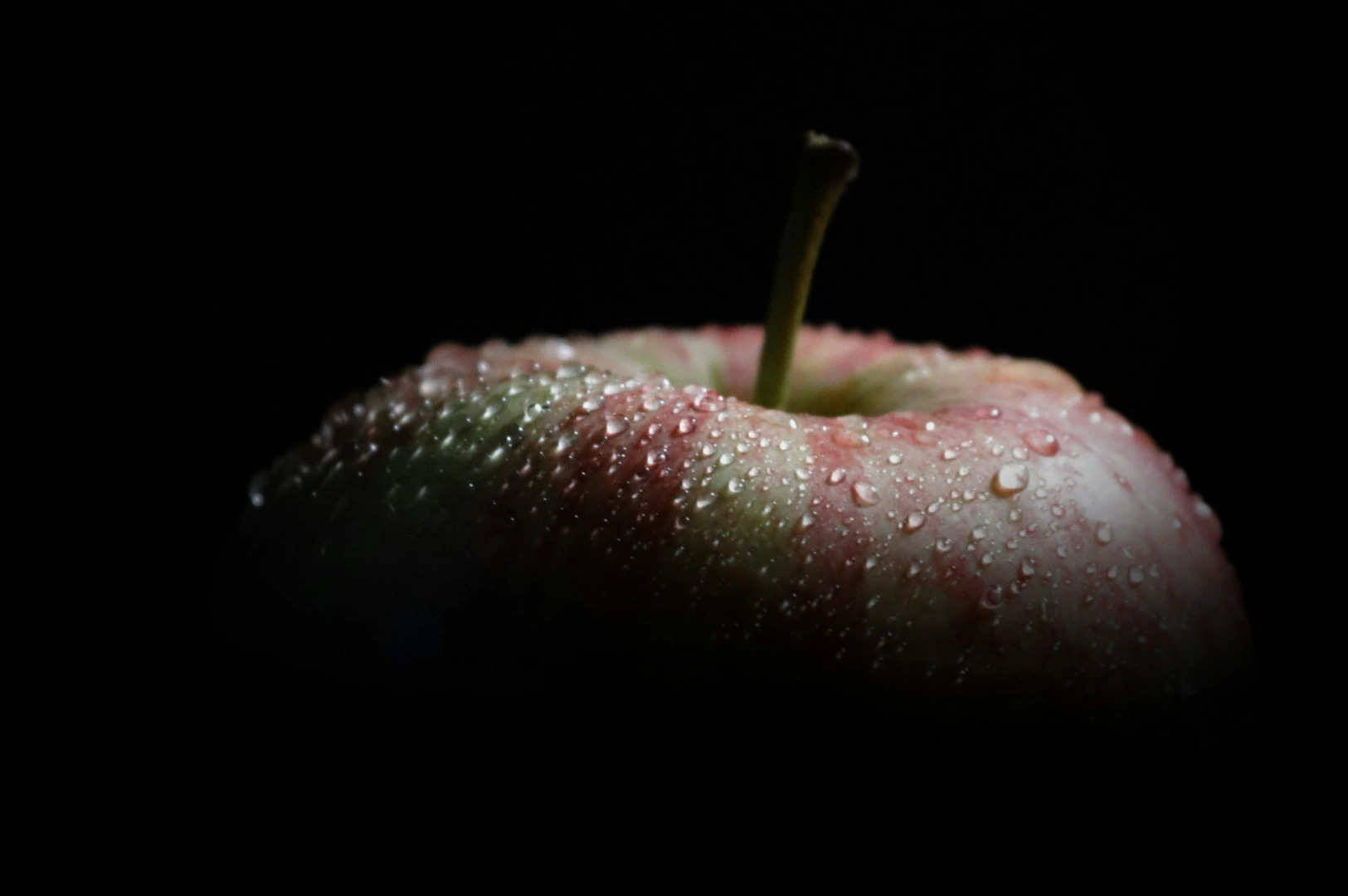 “Close-up of a red apple with water droplets, set against a dark background. The apple is partially illuminated, creating a fresh and dramatic contrast.”