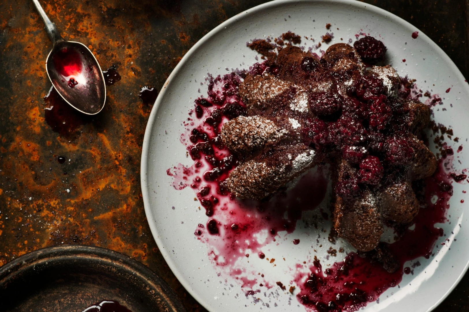 “A chocolate dessert with powdered sugar and vibrant red berries on a white plate, contrasted against a rustic, dark background. A nearby spoon holds berry sauce.”