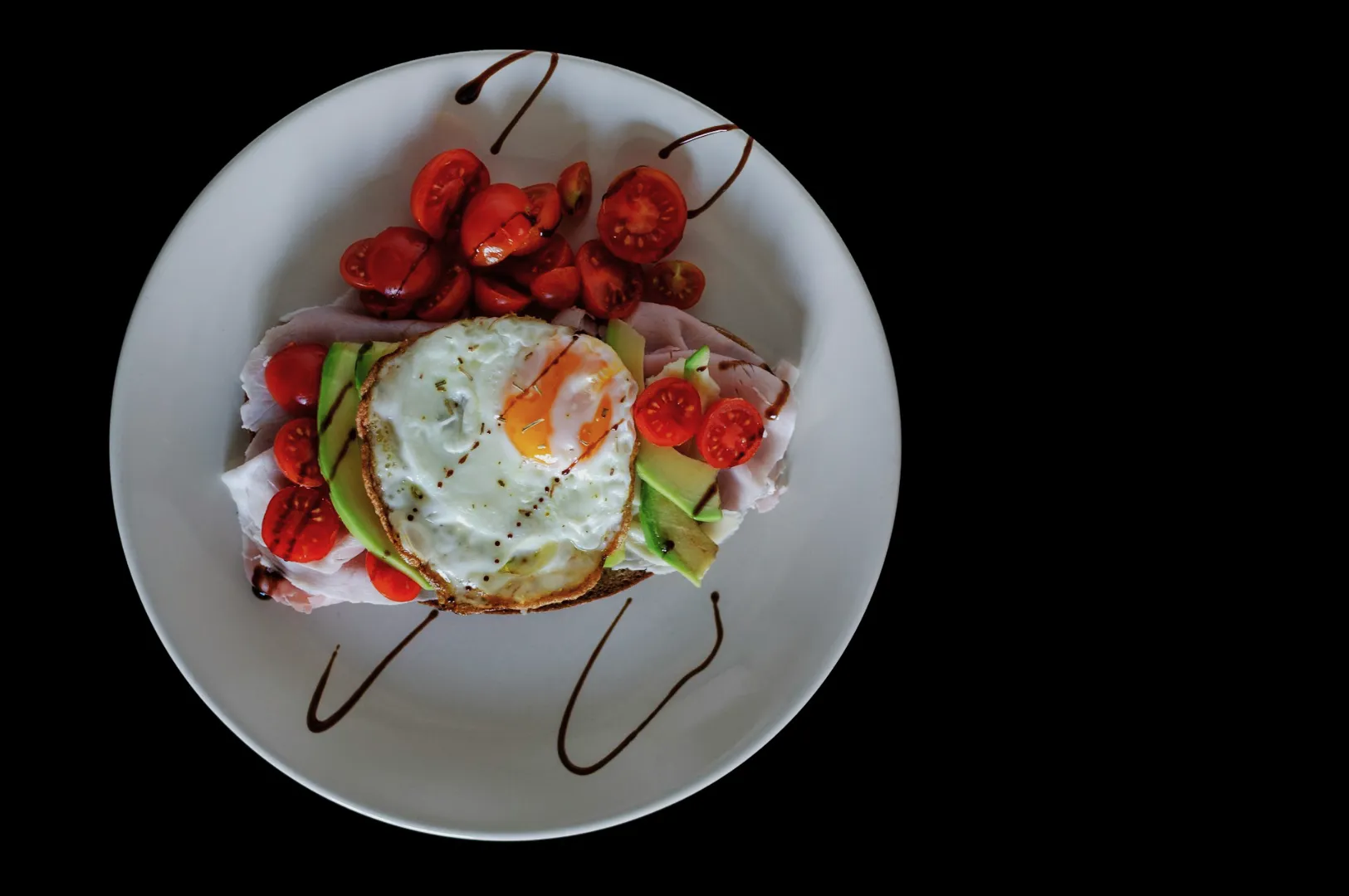 “A white plate holds a slice of bread topped with ham, avocado, and a fried egg. Cherry tomatoes and sauce decorate the plate, all on a black background.”