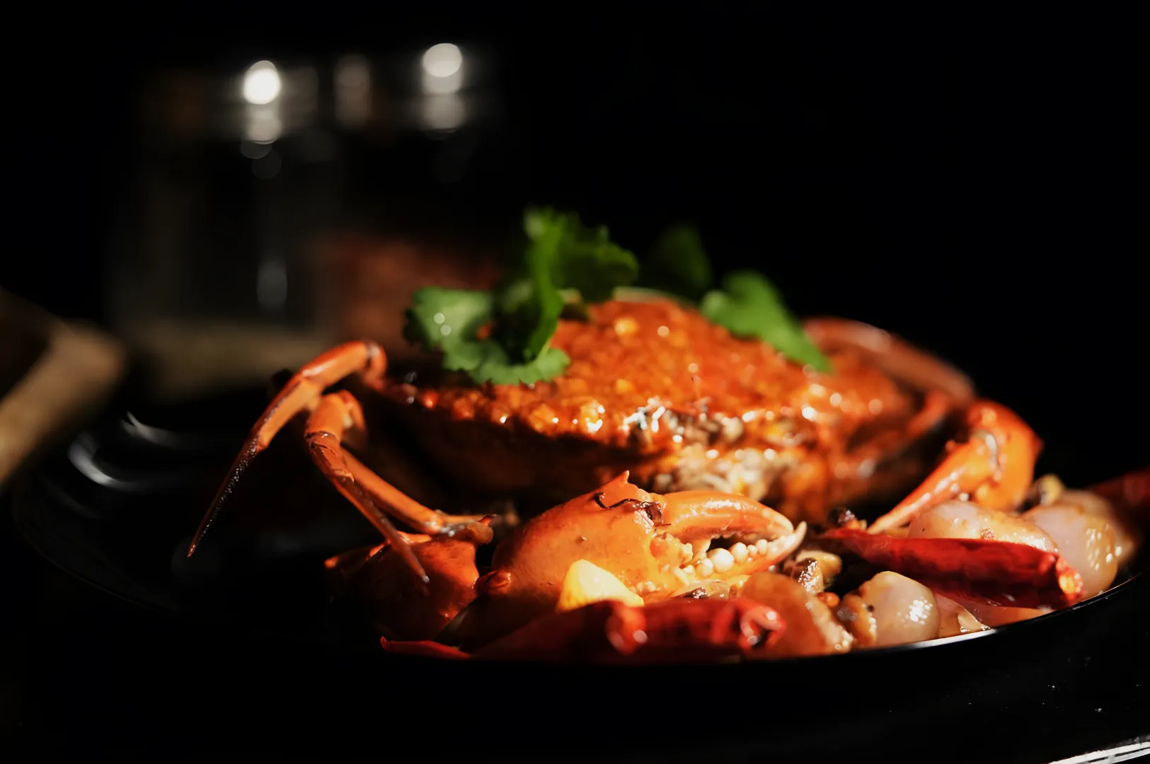 “A chili crab on a black plate garnished with cilantro, surrounded by prawns and dried chili peppers, in dim lighting creating an elegant, appetizing look.”