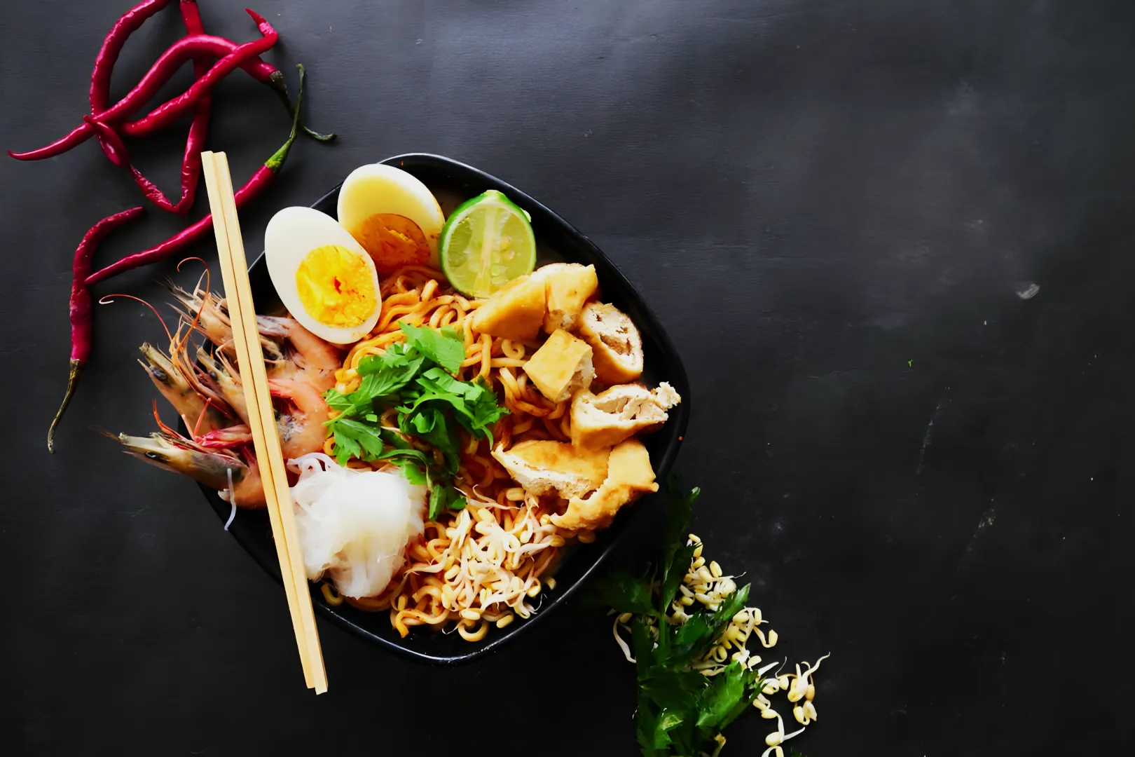 “A black bowl of vibrant noodles garnished with prawns, egg, lime, tofu, and greens sits on a dark surface, with red chili peppers nearby.”
