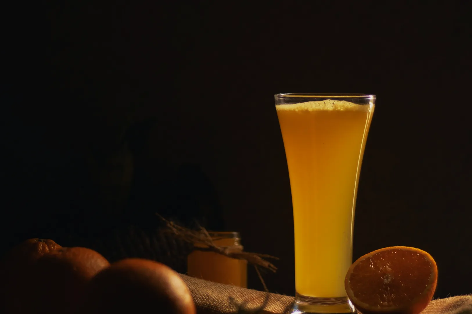 A tall glass of vibrant orange juice sits on a dimly lit table, surrounded by whole oranges and a sliced orange, creating a warm and inviting atmosphere.