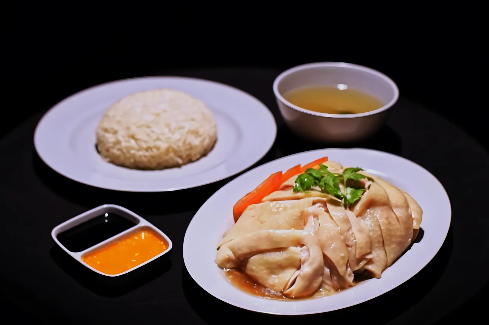 “A plate of sliced poached chicken garnished with cilantro and carrots, beside a serving of rice, clear soup, and dipping sauces, on a dark background.”
