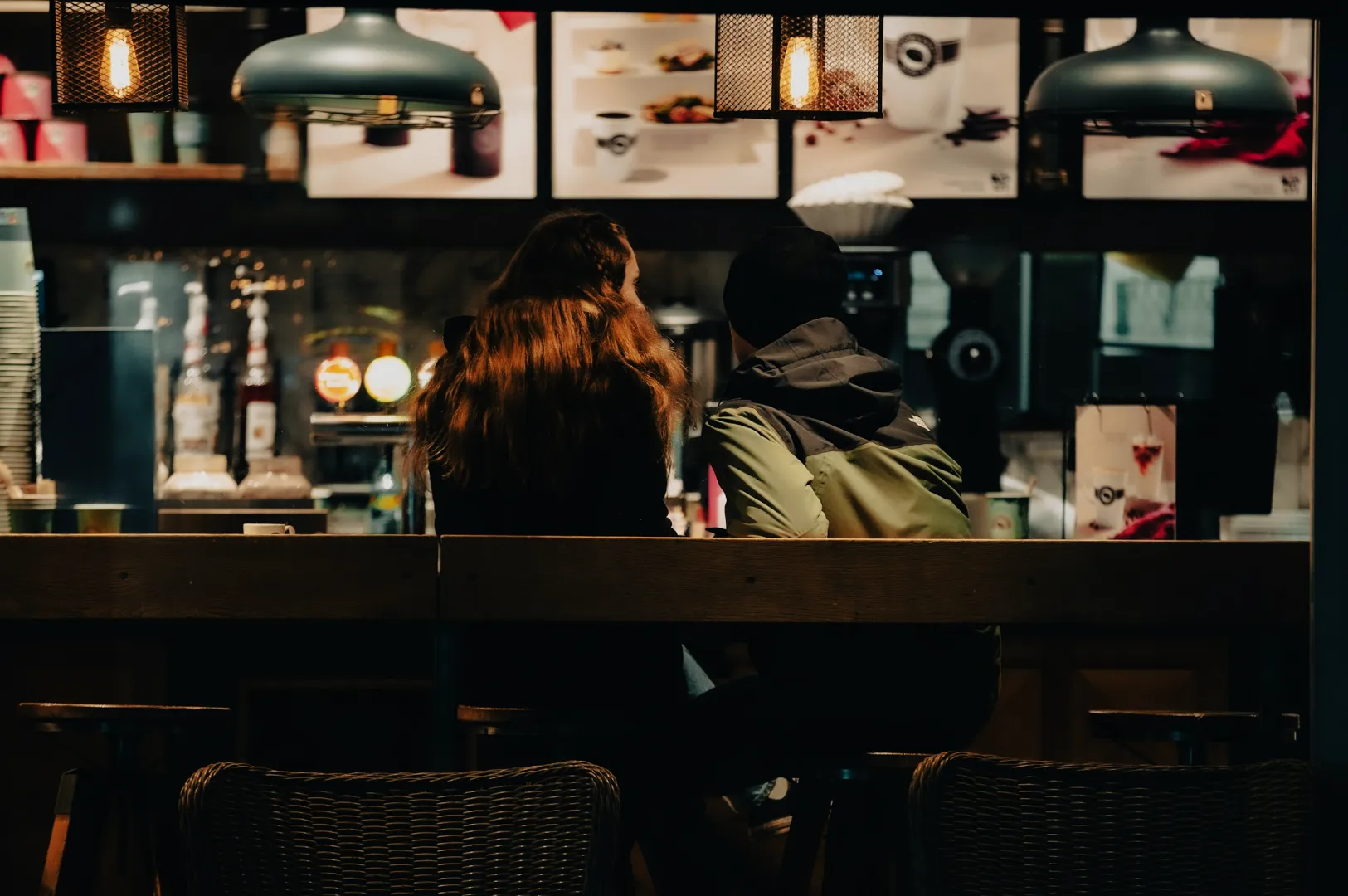 Two people sitting at a dimly lit cafe counter, surrounded by hanging lights and menu boards, creating a cozy and intimate atmosphere.
