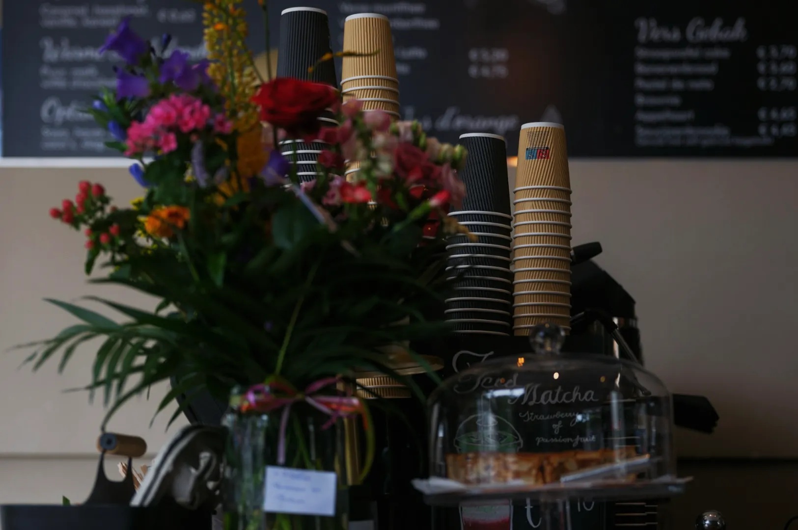 A colorful floral arrangement is in focus with coffee cups stacked in the background. A menu is blurred in the distance, creating a cozy cafe ambiance.