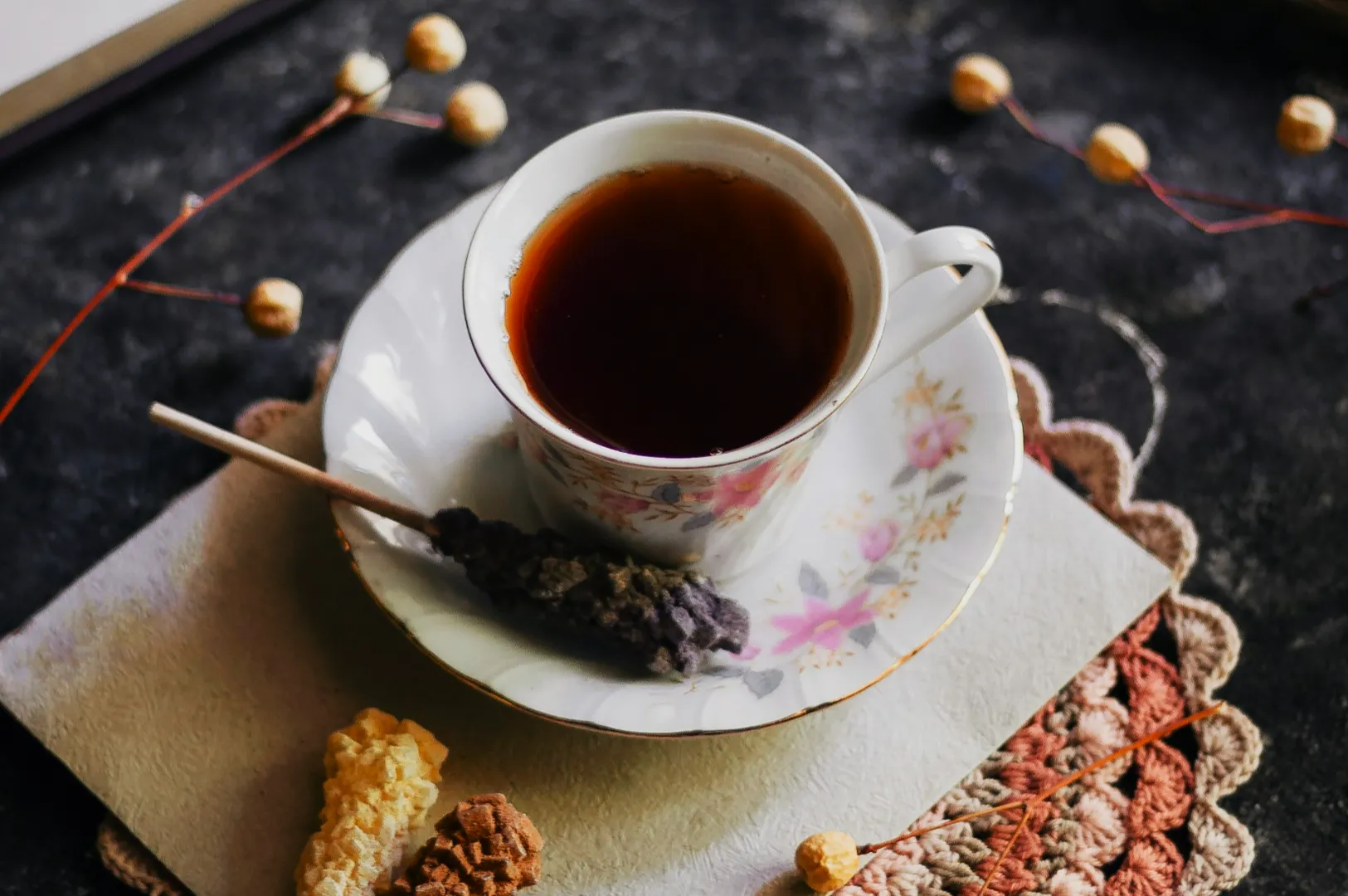 “A floral teacup filled with dark tea sits on a matching saucer. Beside it is a lavender sugar stick, set on a textured placemat, creating a cozy and rustic ambiance.”
