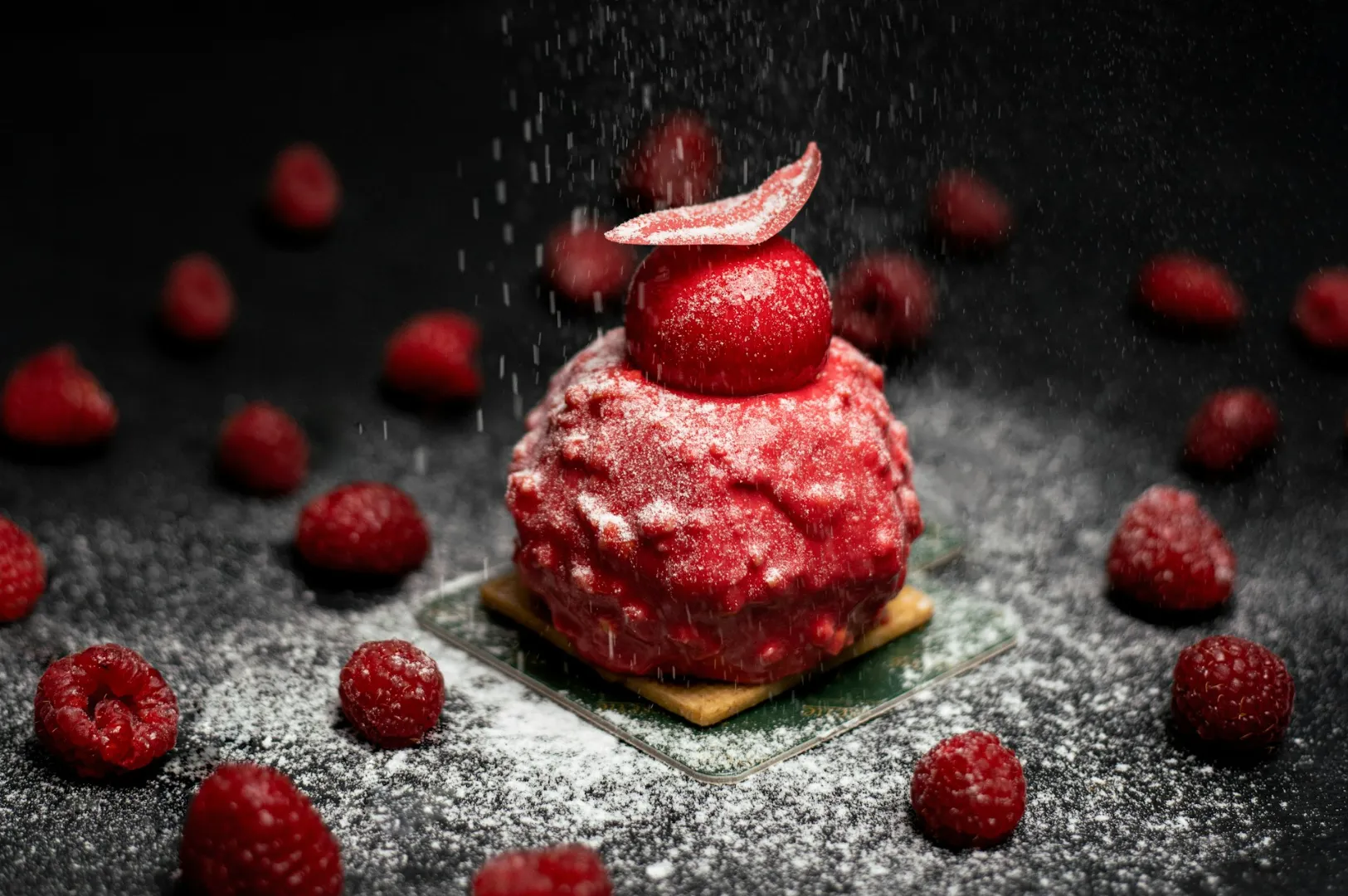 A decorative raspberry dessert sits on a small, square pastry, sprinkled with powdered sugar. Fresh raspberries are scattered on a dark background.