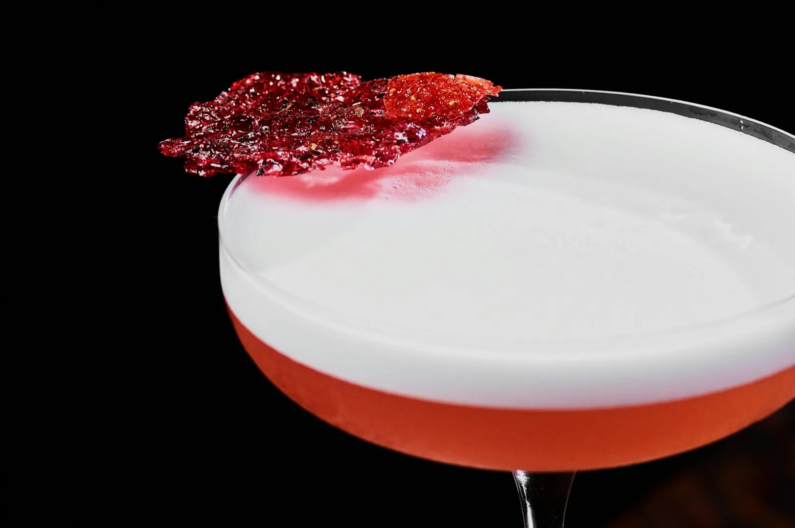 “A close-up of a pink cocktail in a coupe glass with a creamy white foam on top, garnished with a sparkly red sugar shard, set against a black background.”