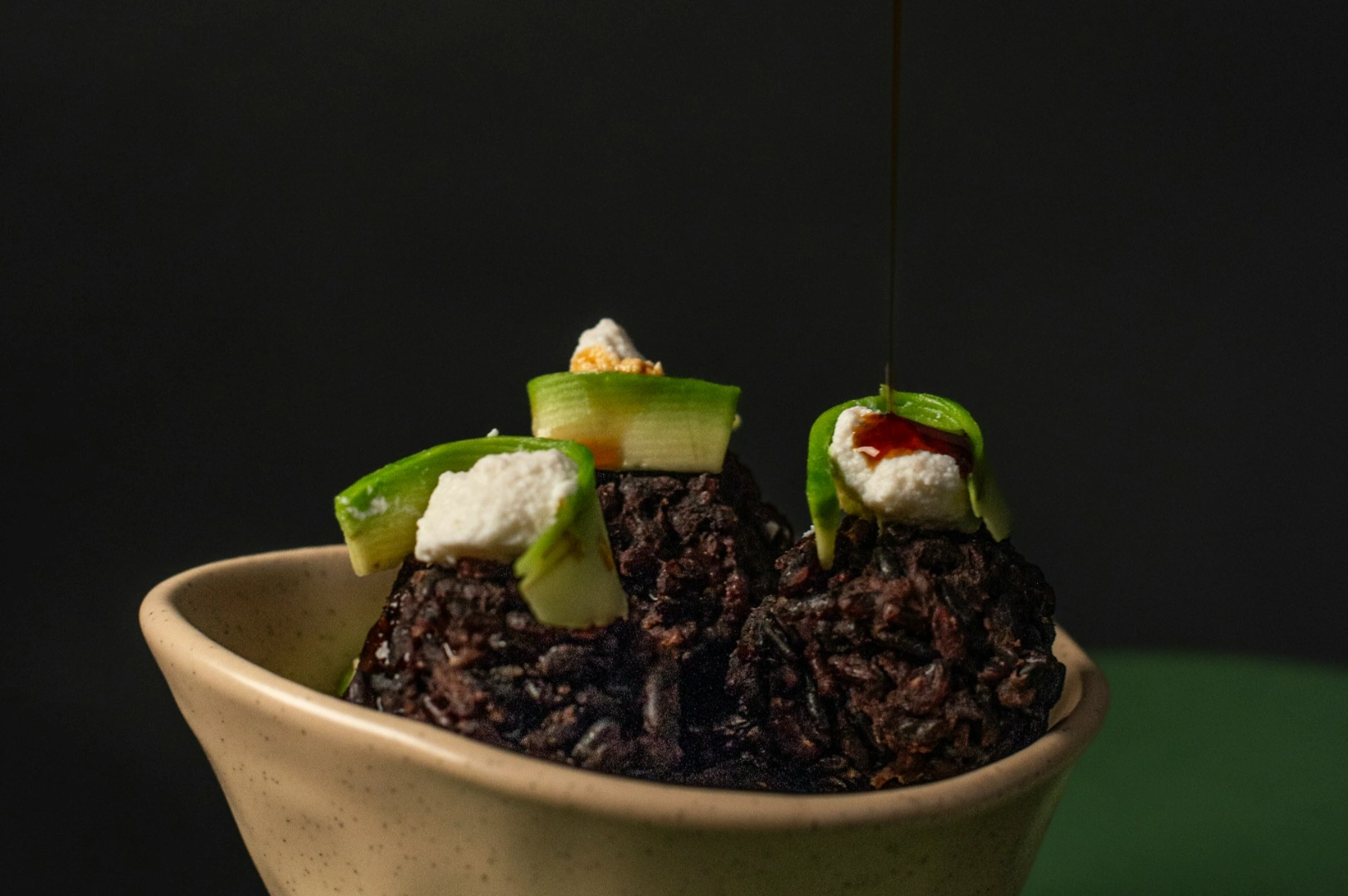 “A beige bowl holds three dark rice balls topped with green strips, white cheese, and sauce. A drizzle of dark liquid falls onto one, set against a dark backdrop.”