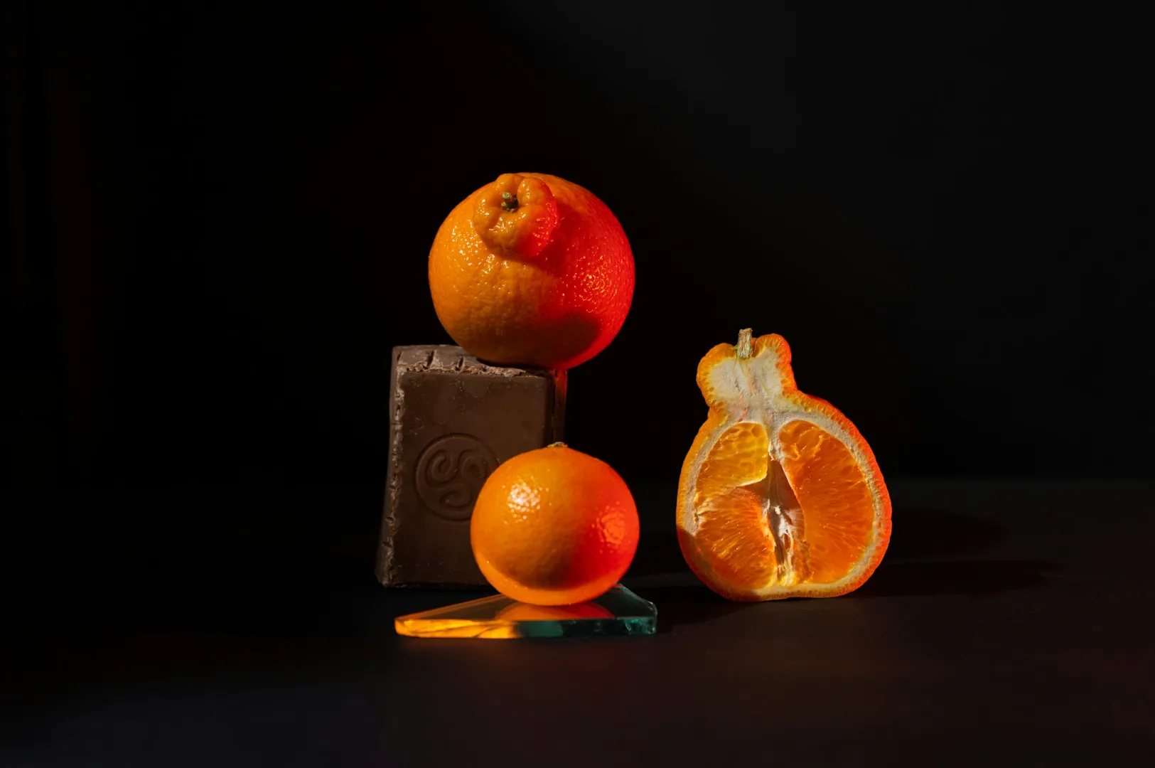 “Three oranges are artfully arranged against a dark background: whole, sliced, and one on a gray block. Warm light creates a dramatic contrast.”