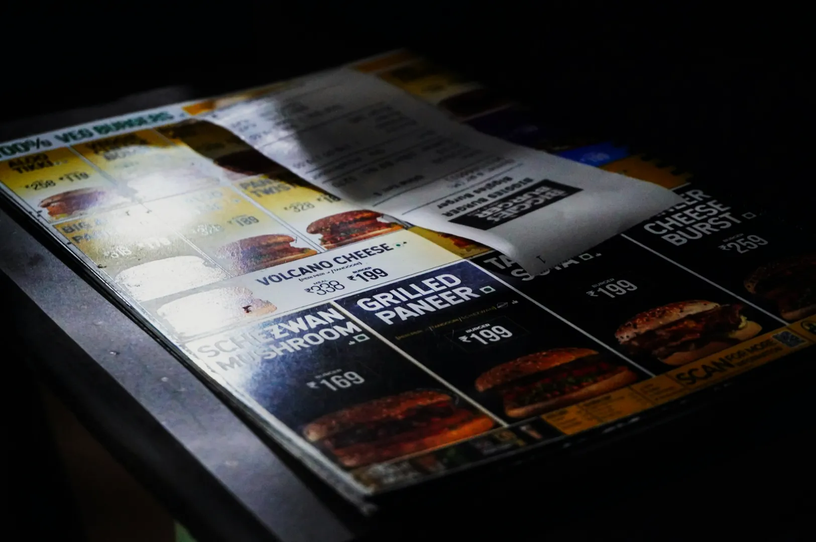 Close-up of a dimly lit menu showing various burger options like "Volcano Cheese" and "Grilled Paneer" with a receipt placed on top.