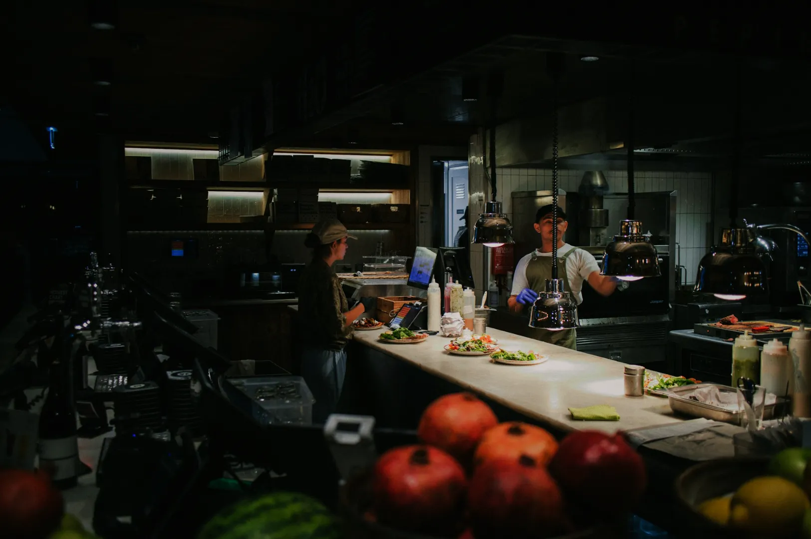 A dimly lit restaurant kitchen with two staff members preparing plates of salad. Fresh pomegranates and other ingredients are visible in the foreground.