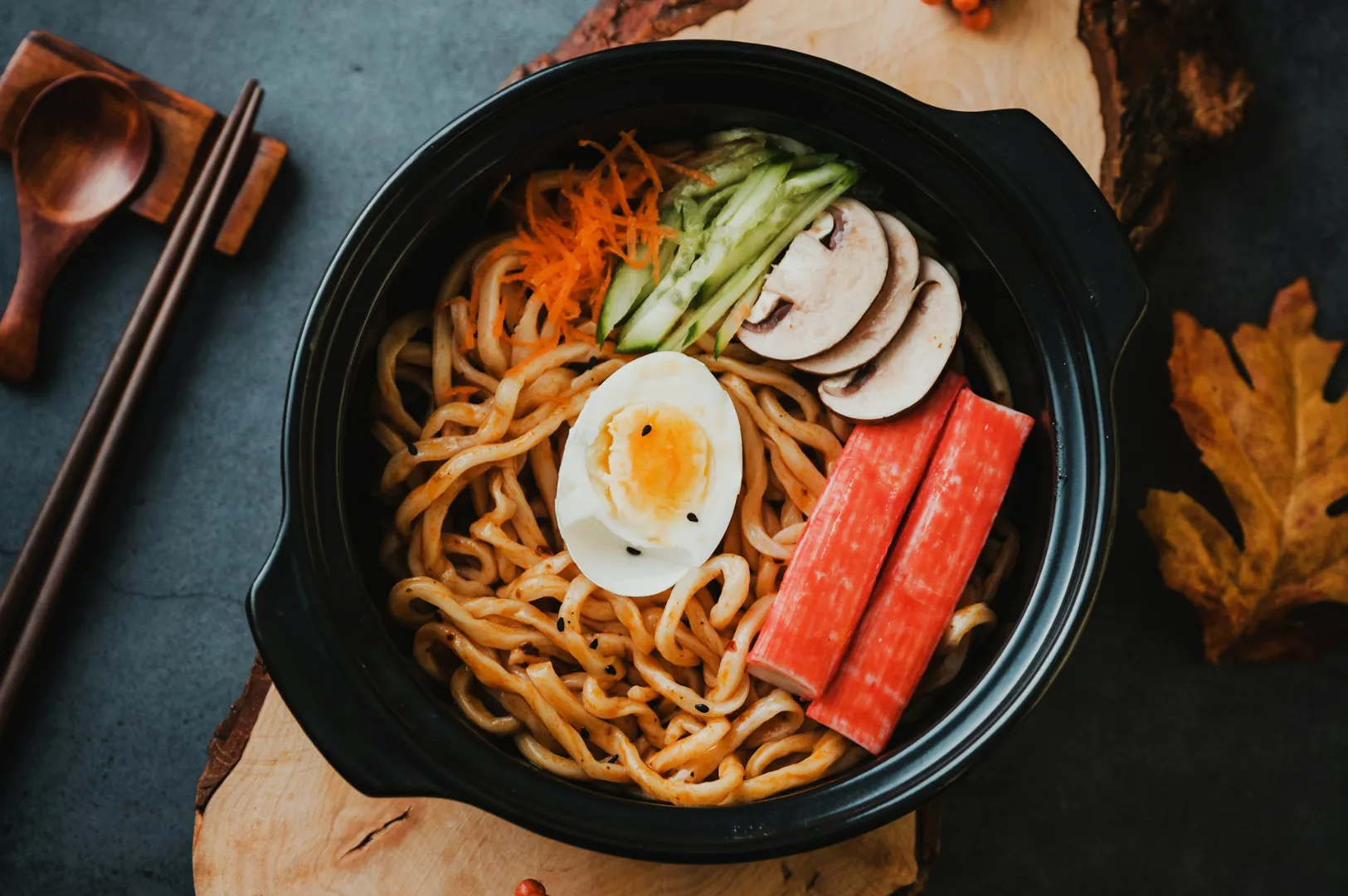 A black bowl of noodles garnished with a halved boiled egg, crab sticks, sliced mushrooms, shredded carrots, and cucumber. Chopsticks and a wooden spoon are nearby.