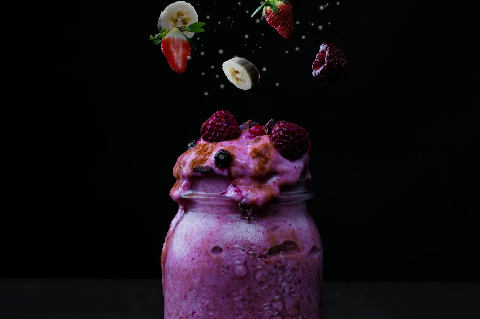 “A mason jar filled with vibrant pink smoothie overflows with raspberries on top. Sliced bananas and strawberries hover above against a black background.”
