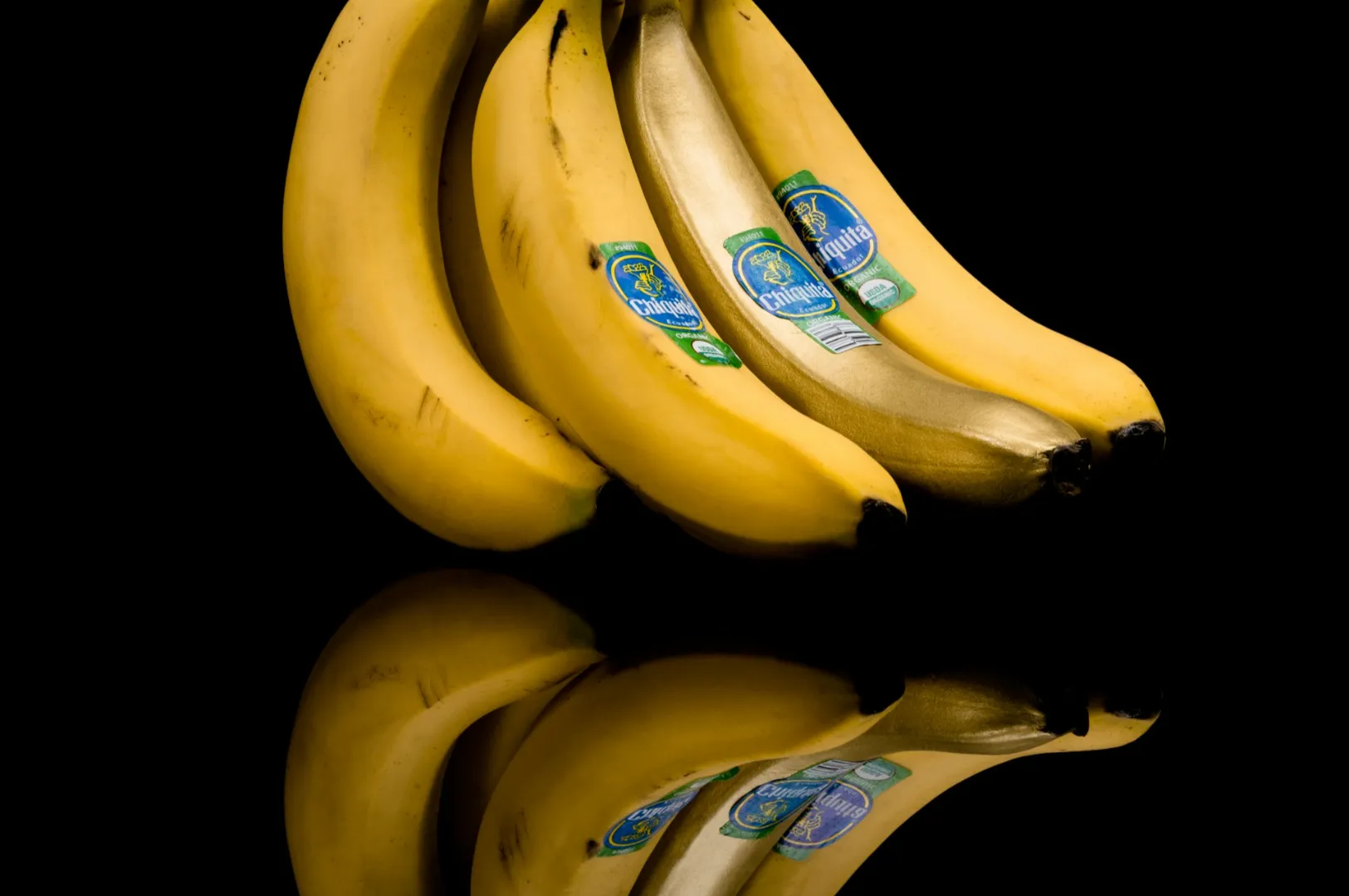 “A bunch of four bright yellow bananas with blue stickers is reflected on a glossy black surface, creating a dramatic and vivid contrast.”
