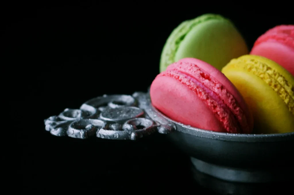 Colorful macarons in pink, yellow, and green are arranged in an ornate silver dish on a dark background, creating a rich and elegant presentation.