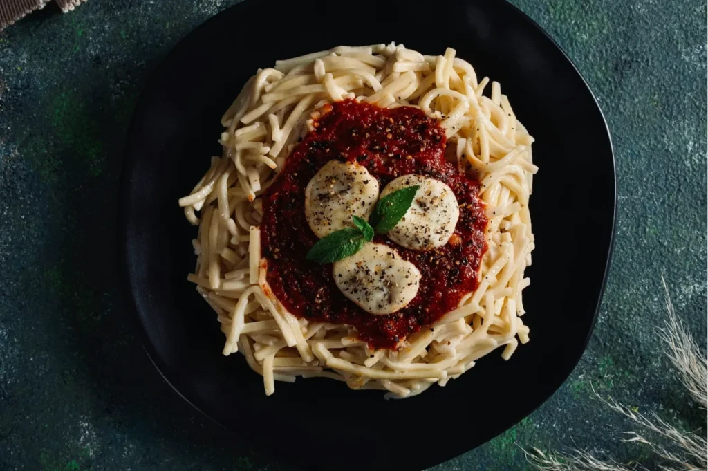 “A black plate with creamy spaghetti, topped with red tomato sauce, three meatballs, and a fresh basil garnish. Set against a dark green textured background.”