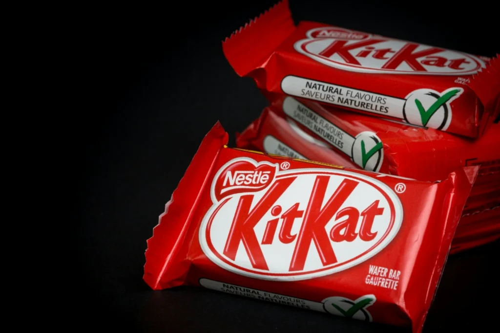 “Red Kit Kat wrapper with bold white and red logo, and green check mark for natural flavors, set against a dark background for contrast.”