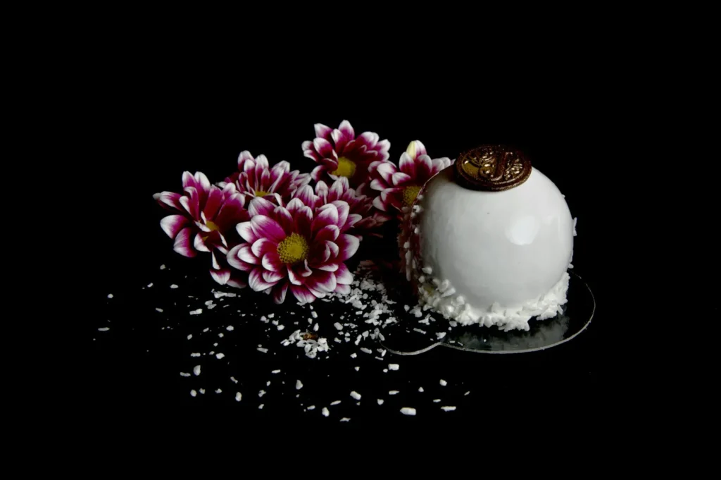“A white, glossy dessert sphere on a black background, garnished with coconut flakes and a chocolate disc, next to vibrant purple and white flowers.”