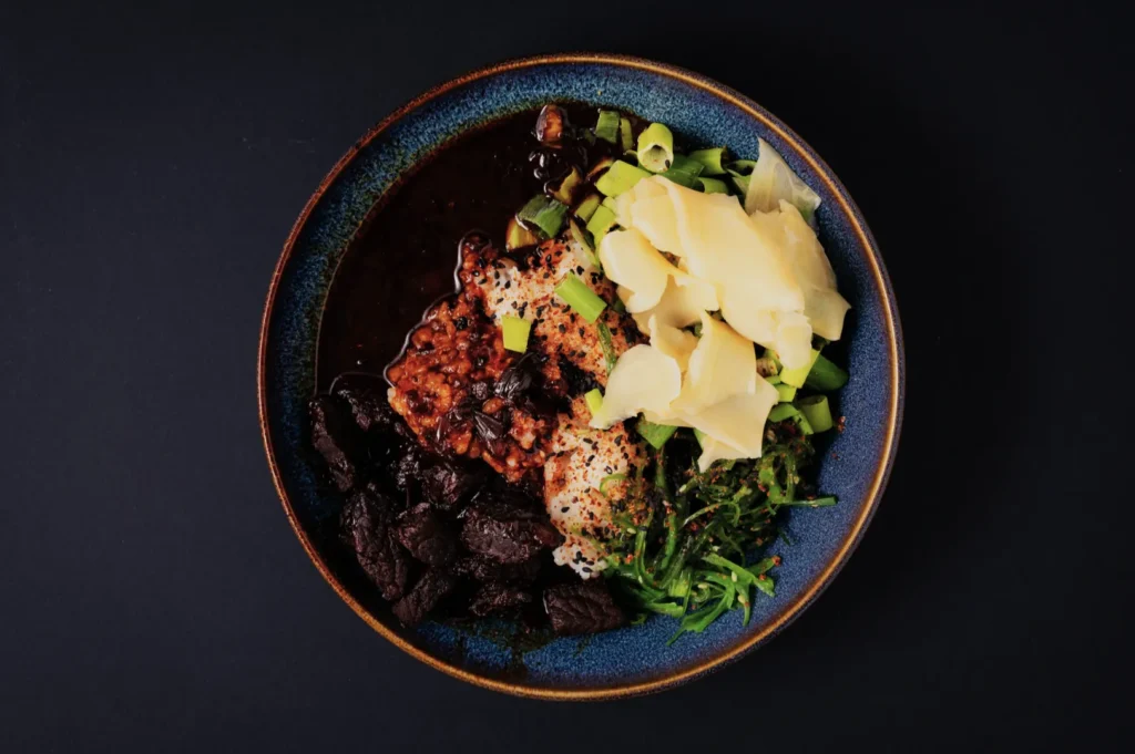 “A vibrant blue bowl on a dark surface, filled with rice topped with black sauce, tender beef slices, green onions, pickled ginger, and seaweed.”