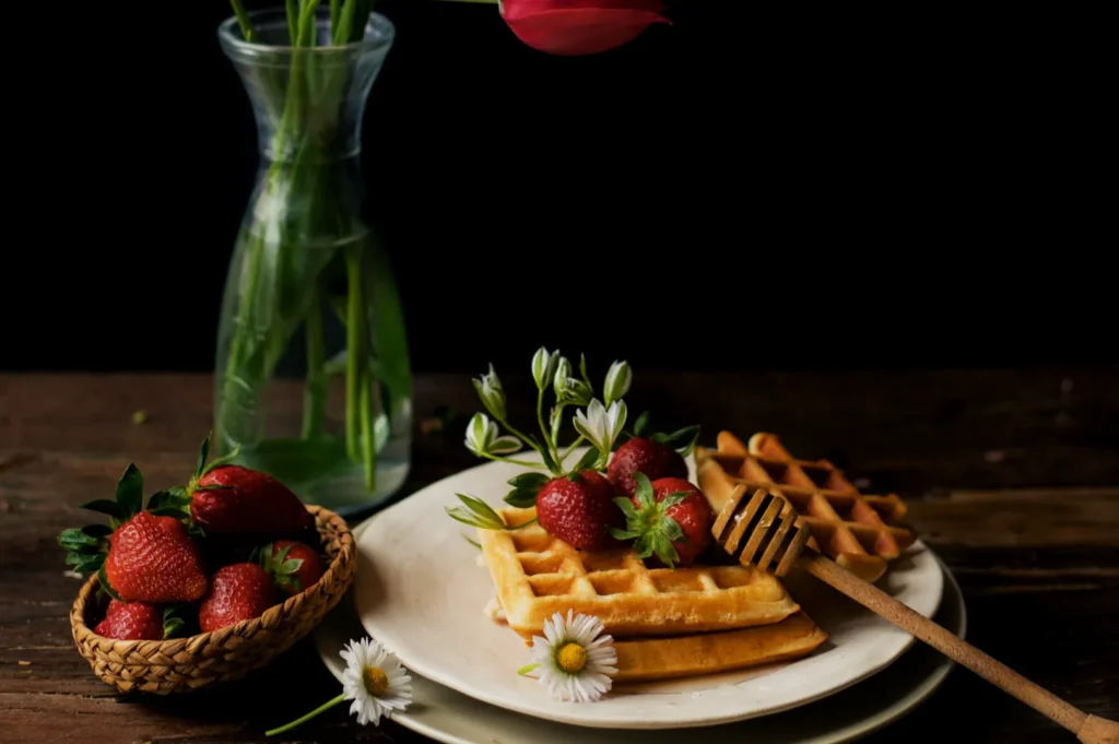 “A rustic setup featuring golden waffles topped with fresh strawberries and white flowers on a plate, beside a honey dipper. A basket of strawberries and a green vase with a single pink tulip complete the scene, set against a dark background.”