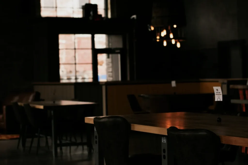 Dimly lit cafe interior with wooden tables and dark chairs, featuring an industrial style chandelier. Large window softly illuminates the room, creating a cozy ambiance.