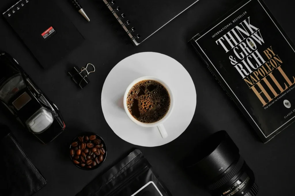 “A flat lay of a white coffee cup on a black table, surrounded by a book, camera lens, pen, notebooks, coffee beans, and a toy car, creating a sophisticated mood.”