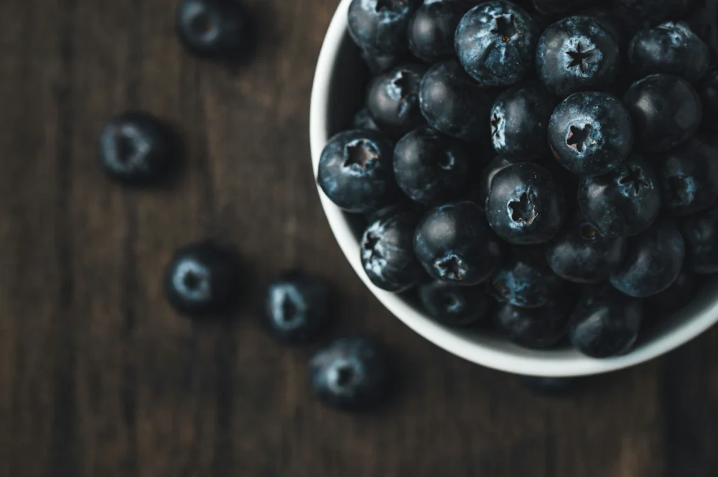 “A bowl filled with fresh blueberries sits on a rustic wooden table. Some berries are scattered around, creating a natural, cozy atmosphere.”