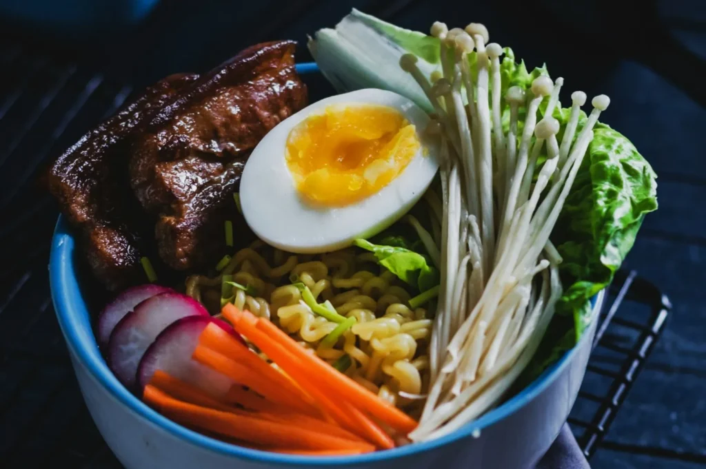 “A bowl of ramen topped with a halved soft-boiled egg, sliced pork, enoki mushrooms, crisp lettuce, radish, and carrot sticks on a dark surface.”