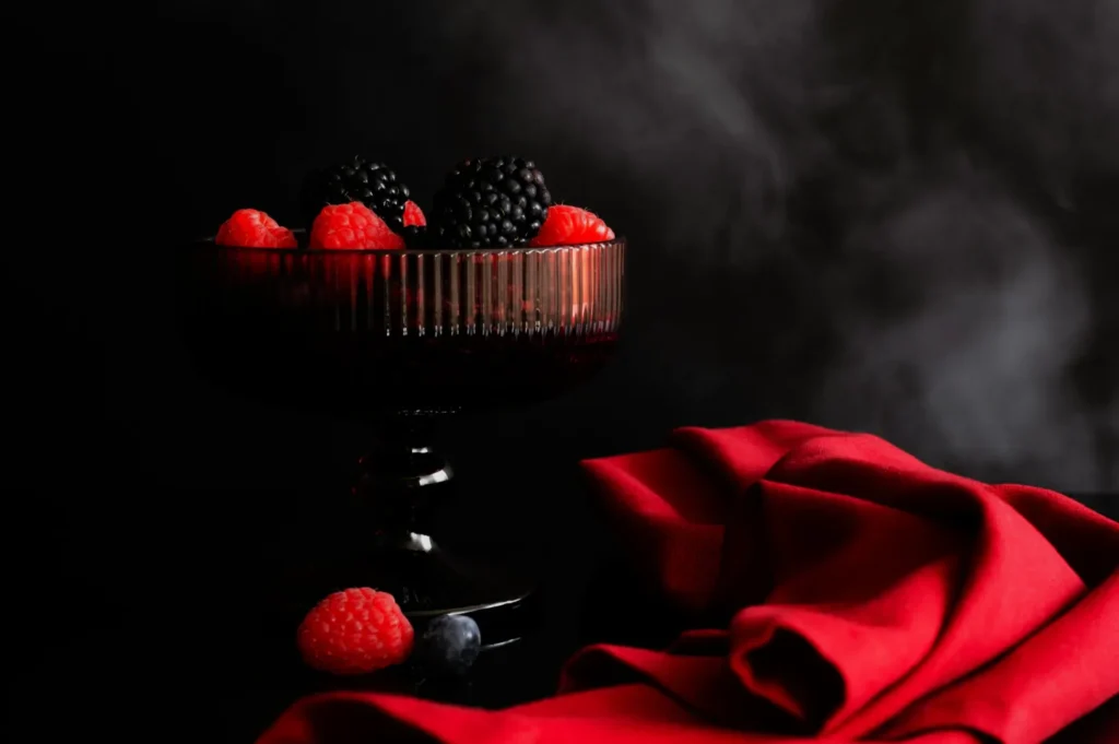 “A dark glass bowl holds vibrant blackberries and raspberries, set against a smoky black backdrop. A rich red cloth adds elegance to the still life.”