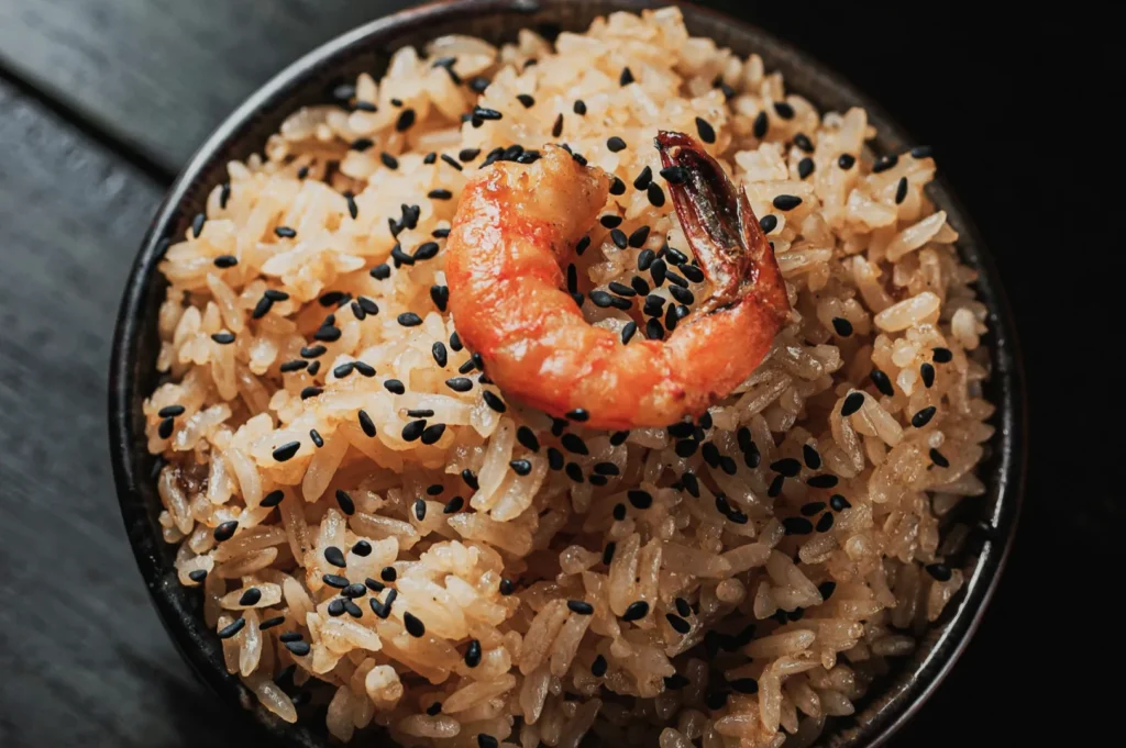 “A bowl of sticky rice topped with a single cooked shrimp and sprinkled with black sesame seeds. The dish appears appetizing and savory.”