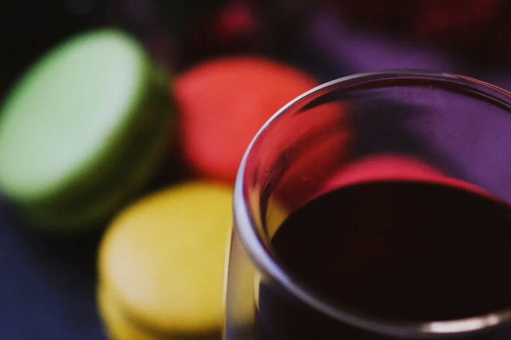“A glass of red wine is in the foreground, with colorful macarons in green, orange, and yellow blurred in the background, creating a cozy, indulgent atmosphere.”