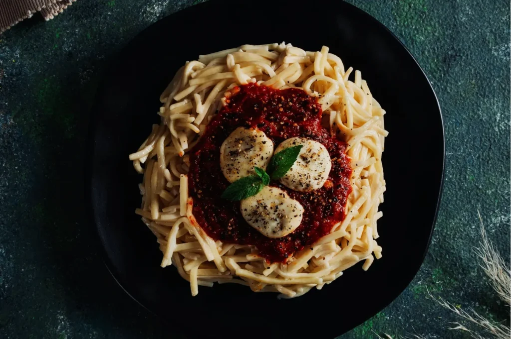 A black plate holds creamy pasta topped with rich tomato sauce and mozzarella slices, garnished with fresh basil, against a rustic dark green backdrop.