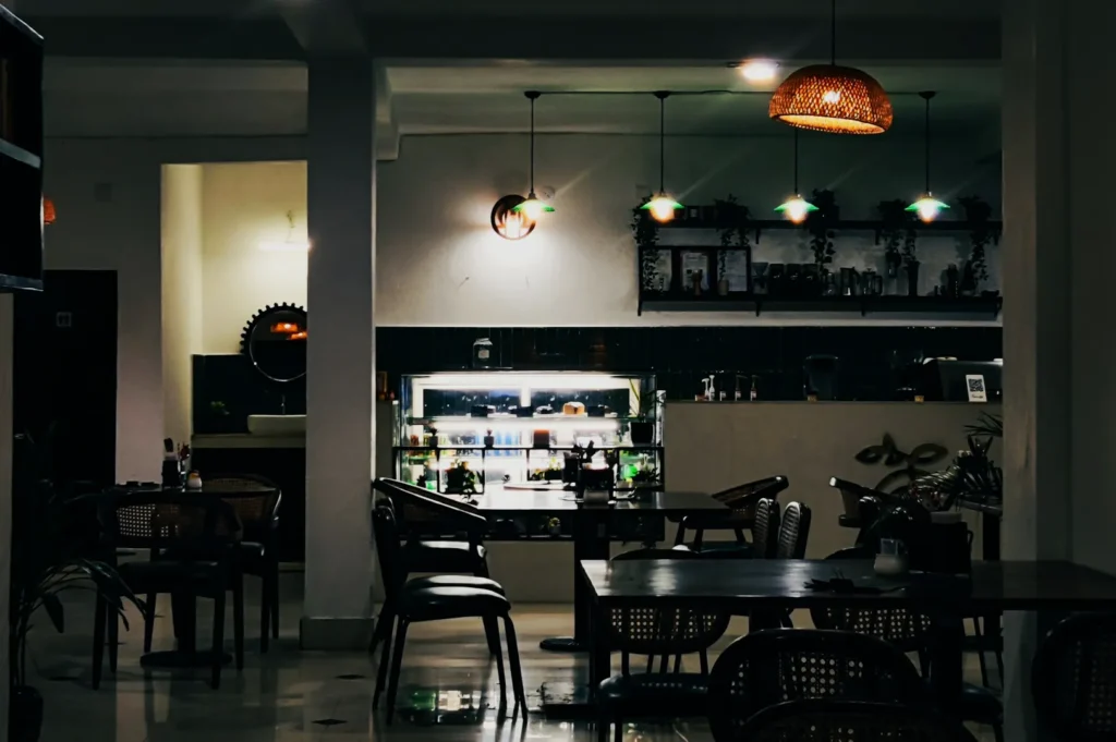 Cozy cafe interior with dim lighting, wooden furniture, and a counter showcasing pastries. Warm ambiance with hanging lights and decorative plants.