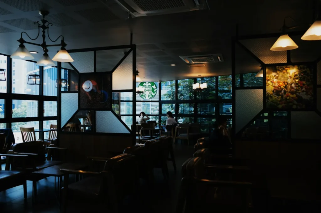 Cozy café with dim lighting, wooden furniture, and decorative wall art. Large windows in the background show a cityscape. A calm, relaxed atmosphere.