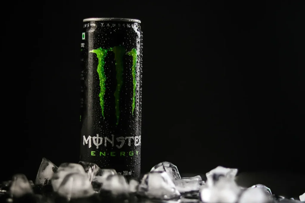 Tall can of Monster Energy drink, covered in condensation, stands against a dark background. Surrounded by scattered ice cubes, it conveys a cool, refreshing feel.