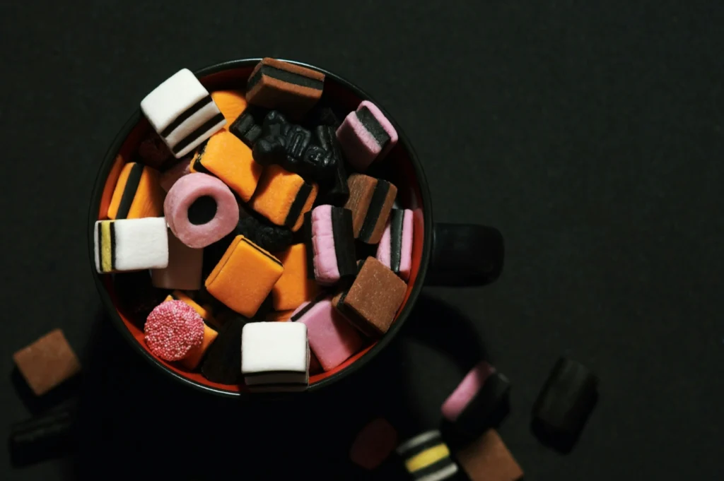 “A black mug filled with colorful licorice allsorts, including orange, pink, and white pieces, scattered on a dark surface, creating a playful vibe.”