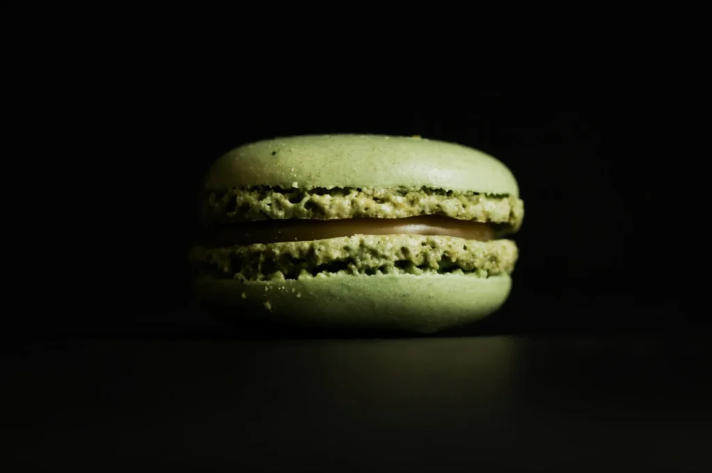 “A single green macaron with textured ridges and a smooth filling sits centered against a dark background. The lighting highlights its delicate structure.”