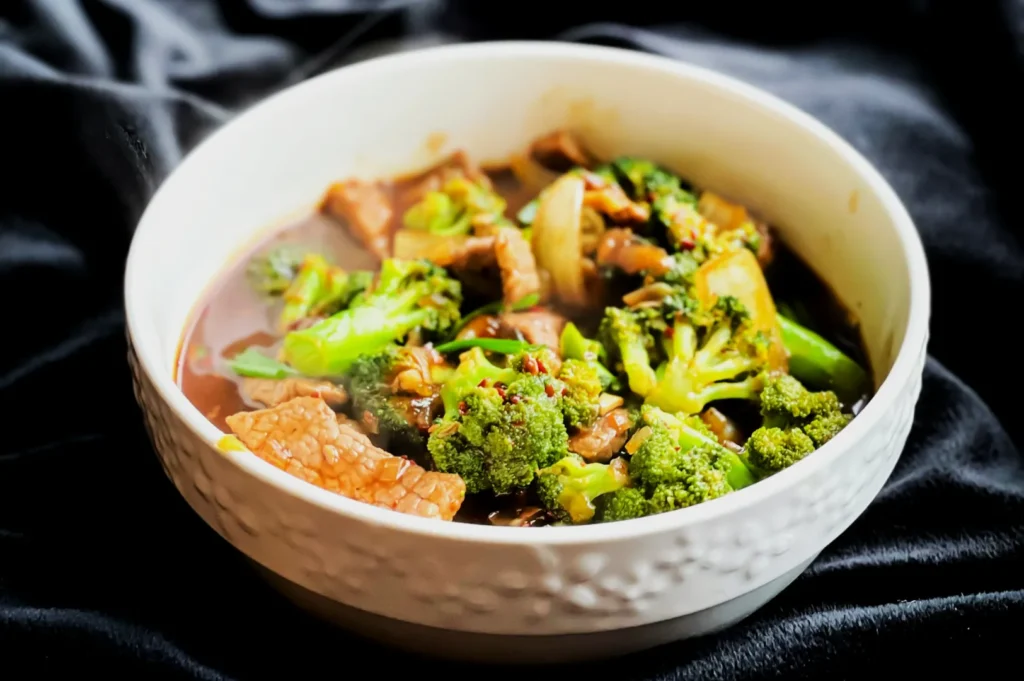 “A white bowl of beef and broccoli stir-fry on a dark cloth background. The dish includes vibrant green broccoli florets and brown beef slices in sauce.”