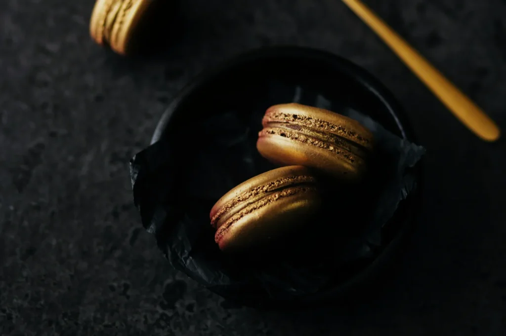 Golden macarons rest on dark paper in a black bowl, set on a textured dark surface. A golden spoon lies nearby, creating an elegant, luxurious mood.