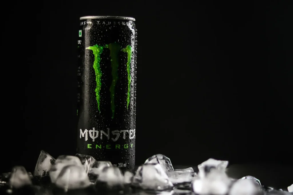 “A can of Monster Energy drink stands upright against a dark background, covered in water droplets, surrounded by scattered ice cubes, conveying a refreshing vibe.”