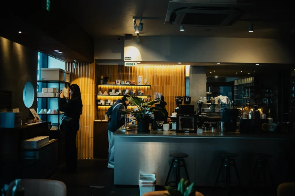 A cozy, dimly lit café features a modern counter with bar stools, a variety of coffee equipment, and plants. Two customers browse and order, creating a warm, inviting atmosphere.