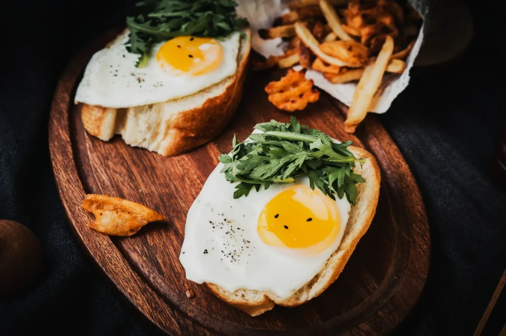 Two slices of bread topped with sunny-side-up eggs and greens on a wooden board, with a side of waffle fries and regular fries. Cozy, rustic setting.