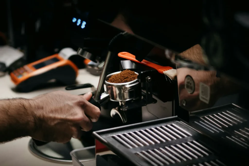 “A person prepares an espresso by tamping coffee grounds in a portafilter, attached to a sleek espresso machine, creating a focused and professional atmosphere.”