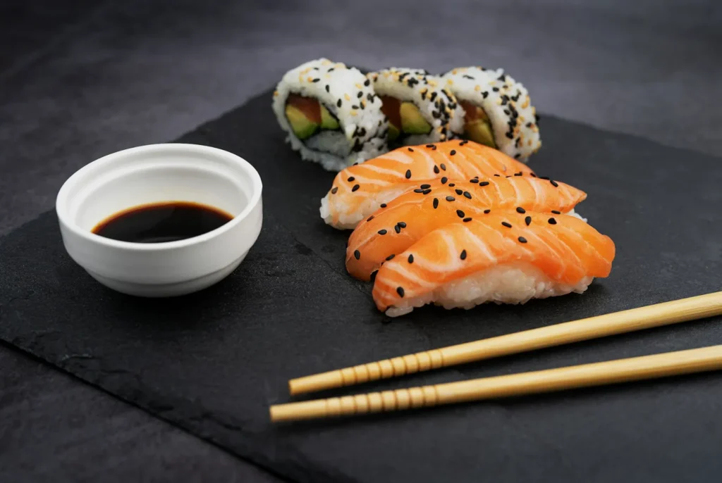 Sushi platter with salmon nigiri and avocado rolls on a slate board, accompanied by soy sauce and wooden chopsticks, creating a fresh, elegant presentation.