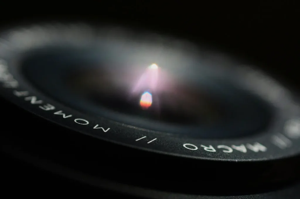 “Close-up of a camera lens with the words “MOMENT MACRO” visible. Soft light reflects off the glass, creating a bright, focused highlight at the center.”