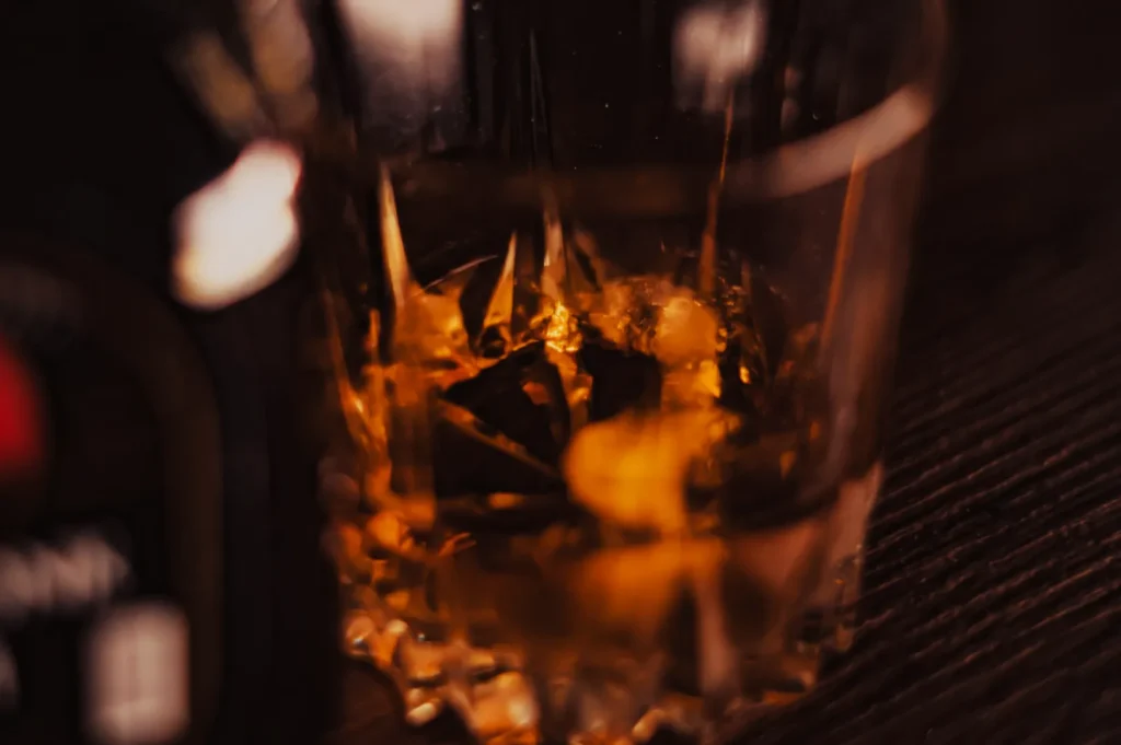 “A close-up of a whiskey glass with ice, casting warm amber tones. The dim lighting and blurred background create a cozy, relaxed atmosphere.”