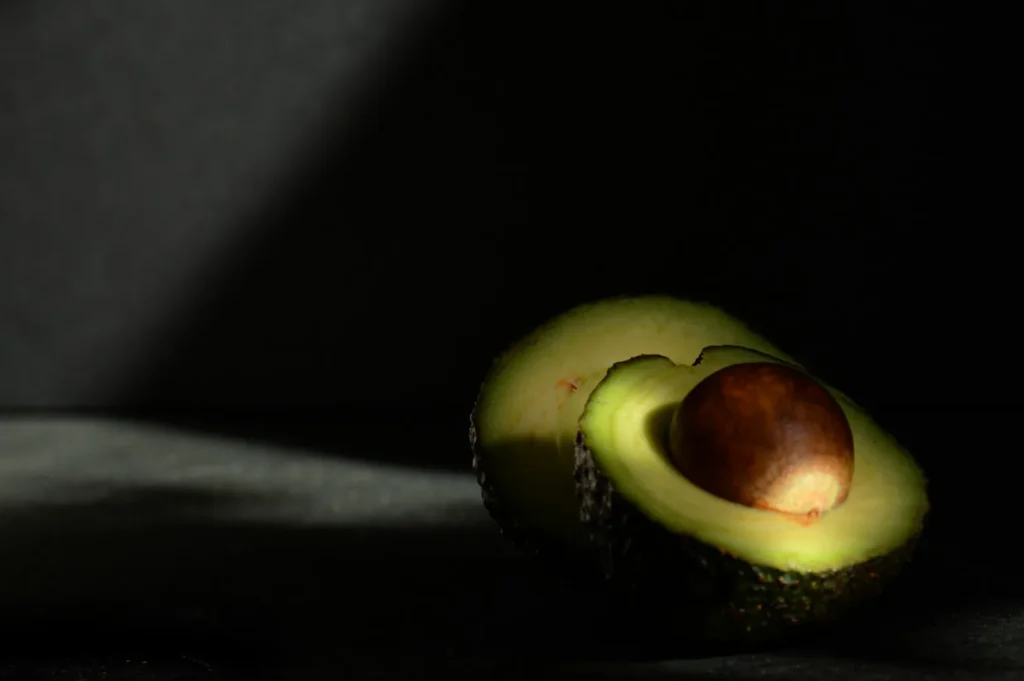 “A halved avocado with its seed is spotlit on a dark surface and background. The lighting casts a dramatic, shadowy contrast, emphasizing its rich texture.”